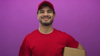 Genç İspanyol adam kırmızı başlık takmış ve mor stüdyo ortamında gülümserken karton kutu tutuyor. Mutluluk..