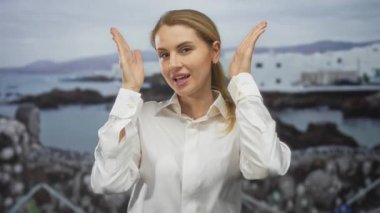 Sahilde poz veren, arka planda beyaz binalar olan beyaz gömlekli, gülümseyen ve açık deniz atmosferini kucaklayan bir kadın..