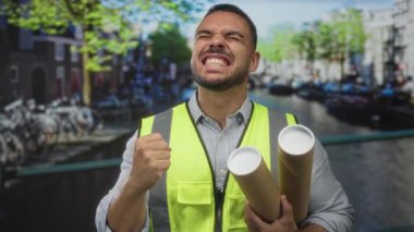 Kanal caddesinde planlar tutarken yansıtıcı yelek giyen genç İspanyol bir adam yumruğunu sıkıyor..