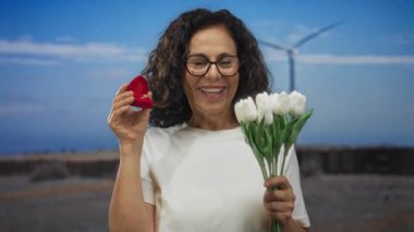 İspanyol kadın neşeyle elinde bir buket beyaz çiçek ve nişan yüzüğü tutuyor dingin bir arka planda yel değirmeninin yanında, samimi bir açık hava evlenme teklifi anını yakalıyor..