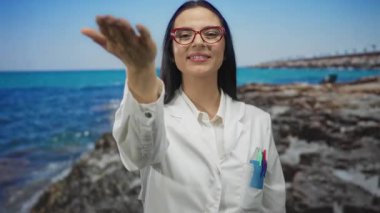 Üniformalı İspanyol kadın bilim adamı deniz kenarındaki plajda elini uzatarak canlı bir kıyı arkaplanı ve profesyonel giysi sergiliyor..