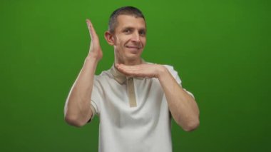 Kafkasyalı bir adam neşeli bir şekilde gülümsüyor. Parlak yeşil arka planda el kol hareketi yapıyor. Neşeli bir yüz ifadesi var..