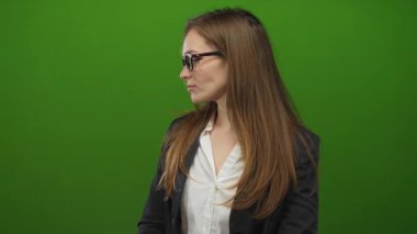 Uzun kahverengi saçlı, gözlüklü ve spor ceketli genç bir kadın yeşil stüdyoda ağzını açar..