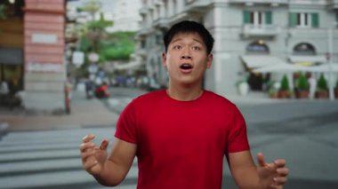 Kırmızı gömlekli genç adam şehir sokaklarında heyecan gösteriyor. Geçmişi bulanık, şehir enerjisini yakalıyor ve yaşam tarzı görüntüsü için dinamik bir ifade oluşturuyor.