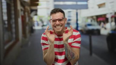 Gözlüklü ve kolunda dövmesi olan genç adam şehir sokaklarında çizgili gömlekle duygularını ifade ediyor..