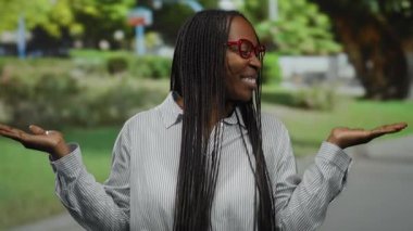 Açık hava parkında gülümseyen, yemyeşil ağaçlar ve bulanık bir arka planı olan, gözlüklü ve çizgili gömlekli, kollarını kaldırmış, neşeli bir jest yapan bir kadın..