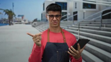 Genç adam güneşli bir günde önlük giyer, tablet kullanır. Arka planda şaşkın, şehirli gibi görünür. Canlı ve günlük giysiler giyer..