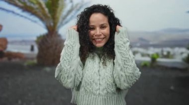 Parkta gülümseyen bir kadın palmiye ağaçlarının altında rahat bir süveter giyiyor, gündüz doğal ortamında neşeli bir ifade sergiliyor..