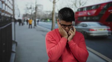 Kırmızı kazaklı genç adam arka planda kırmızı çift katlı otobüslerle şehir caddesinde telefonla konuşuyor.