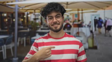 Gözlüklü ve çizgili gömlekli genç sakallı adam gülümsüyor ve arka planda kafesi olan şehir sokaklarında kendini işaret ediyor..