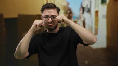 Sakallı ve gözlüklü genç bir adam, şehir sokaklarında ağlıyormuş gibi yapıyor oyuncu ifadeler ve komik duygular sergiliyor..
