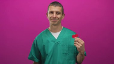 Yeşil üniformalı adam kırmızı kalbi canlı pembe bir arka plana karşı tutarak sıcak ve kendine güvenen neşeli bir tıp uzmanını gösteriyor..
