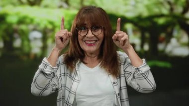 Kıdemli İspanyol kadın canlı bir açık hava parkı ortamında gülümsüyor, iki parmağıyla yukarıya doğru işaret ediyor, yemyeşil bir zemin arasında mutluluk ve pozitiflik taşıyor.