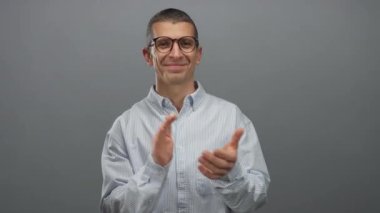 Gözlüklü ve çizgili tişörtlü bir adam izole edilmiş gri duvar arkaplanına karşı alkış tutuyor. Onayını ve pozitifliğini çağdaş bir ortamda, kendine güvenen bir gülümsemeyle ifade ediyor.