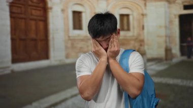 Şehir caddesinde mavi bir sırt çantasıyla gezen genç adam stresli görünüyor, dış mekan ayarları bir öğrencinin şehir ortamını vurgulayarak zorluklarla ya da duygularla karşı karşıya olduğunu gösteriyor..