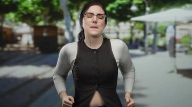 Kumral saçlı bir kadın, açık havada, güneşli bir sokakta, yeşil ve kentsel manzarayla çevrili gözlük ve günlük giysiler takarak duygularını ifade ediyor..