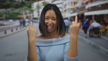 Çinli kadın şakacı bir şekilde dilini dışarı çıkarıyor ve arka planda yayalar ve binalar olan kalabalık bir şehir caddesinde el kol geziyor..