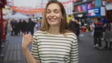 Çizgili kazak giyen bir kadın, kalabalık bir şehir caddesi pazarında, önünden geçen bulanık yayalarla baş parmak işareti yapıyor..