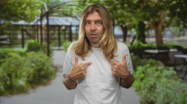 Uzun sarı saçlı ve beyaz gömlekli bir adam yeşil çalılar ve boş banklarla kaplı dar orman yolunda başparmaklarını göğsüne uzatıyor. Kendinden şüphe ediyor..