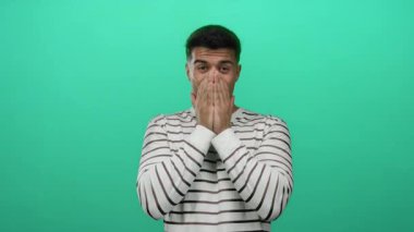 Genç adam elleri ile ağzını kapatıyor yeşil bir zemine karşı neşeli bir şekilde duruyor çizgili bir gömlek giyiyor duygularını ve şaşkınlığını gösteriyor..