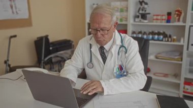 Klinikteki kıdemli erkek doktor yanında ekipman ve ilaç raflarıyla çevrili tıbbi belgelerle bir dizüstü bilgisayar üzerinde çalışıyor..