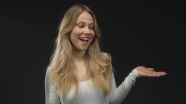 Sarı saçlı ve beyaz yüzlü bir kadın parlak bir şekilde gülümser ve stüdyoda avucunu açmak için işaret eder; merak uyandıran nişan daveti.