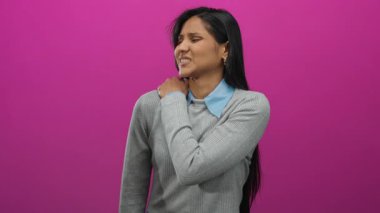 Sırt ağrısı çeken bir kadın omuzlarını canlı pembe bir arka plana karşı rahatsızlıkla tutuyor, genç Latin kültürü ve izolasyon üzerine odaklı bir sağlık hizmeti teması sunuyor..