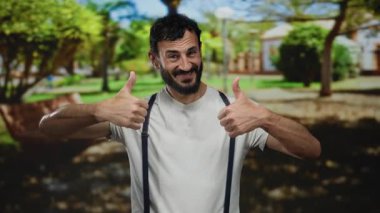 Ağaçlarla çevrili yeşil bir parkta gülümseyen ve baş parmağını kaldıran sakallı bir adam olumlu ve neşeli bir atmosfer olduğunu gösteriyor..