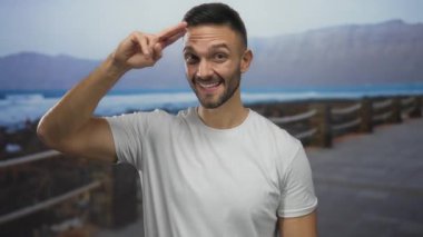 İspanyol genç adam, parmakları kafasında selamlıyor, deniz kenarında gezinti arka planında mavi deniz ve dağlarla gülümsüyor, açık havada günlük beyaz tişört giyiyor..