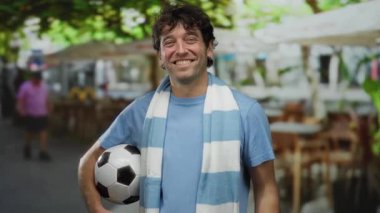 İspanyol bir adam sokakta kutlama yapıyor, elinde bir futbol topu tutuyor ve büyük bir gülümsemeyle parmaklarını sayıyor, spora ve canlı şehir atmosferine olan tutkusunu yakalıyor..