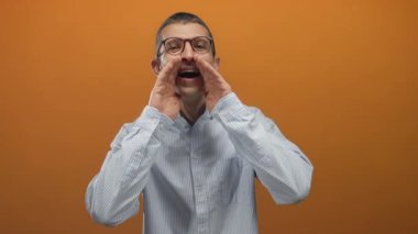 Elleri ağızlarının önünde bağlanmış, gözlüklü ve çizgili gömlekli, izole edilmiş turuncu arka planda bağıran bir adam..
