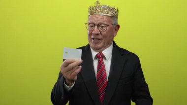 Taç giyen kıdemli bir adam boş bir kredi kartı tutuyor ve izole edilmiş sarı bir arkaplana işaret ediyor. Takım elbise giymiş, güven ve başarıyı ifade ediyor..