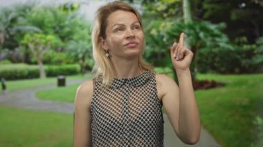 Sarışın kadın geniş bir gülümseme ve yeşil orman ağaçları arasında başparmak uzatmak; iyimserlik onayı.