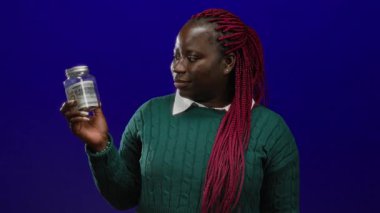 Dolarlık banknot içeren kavanozu canlı mavi arka planda tutan kadın para ve ifade odaklı finansal konsepti yakalıyor.