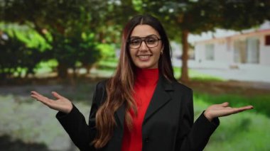 Parkta elleri açık, kırmızı ve siyah bir şekilde gülümseyen bir kadın, genç bir ifade, İspanyol, yeşil gözlüklü, etrafı yeşilliklerle çevrili..