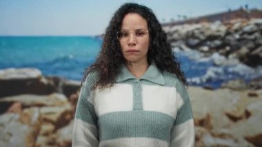 Gözlüklü bir kadın berrak bir gökyüzünün altında parlak mavi bir denizle kayalık bir sahilde duruyor, dingin ama düşünceli bir hava yayıyor..