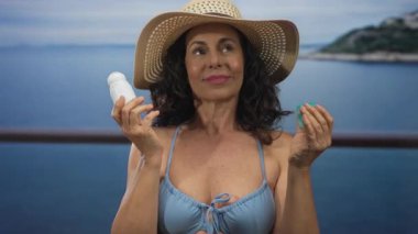 Kadın, elinde bikini ve şapkayla güneşlenirken sakin plaj titreşimlerinden zevk alıyor. Mavi deniz kenarında sakin bir arka planı var. Açık havada bir dinlenme anı yakalıyor..