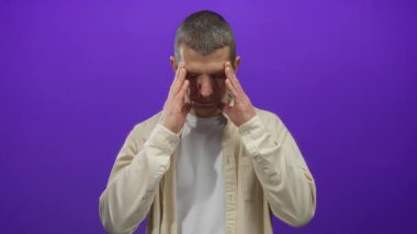 Kısa saçlı, bej gömlekli bir adam, mor bir arka plan duvarına karşı düşünceleriyle şakaklarına dokunur, stres ve düşünce duyguları taşır..