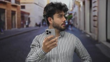 Sakallı genç adam şehir caddesinde akıllı telefon kullanıyor çizgili tişört giyiyor günlük ortamda çevreyi gözetliyor sokak deneyimlerini geliştiriyor..