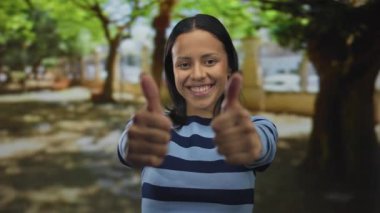 Güneşli bir parkta yeşil ağaçlarla gülümseyen ve açık havada çizgili gömlek giyen İspanyol bir kadın..