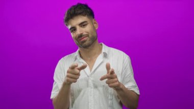 Stüdyoda mor bir arka plana gülümserken, beyaz keten gömlek giyen genç İspanyol adam parmaklarını işaret ediyor..