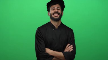 Siyah üniformalı, gülümseyen İspanyol erkek şef canlı yeşil arka planda aşçılık uzmanlığına karşı güvenle duruyor..