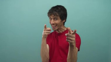 Kırmızı tişörtlü adam kamerayı işaret ediyor. Stüdyoda tepe duvarına karşı geniş bir sırıtışla..