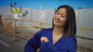 İnşaat alanında barikatlarla gülümseyen, el kol hareketi yapan mavi bluz giyen, açık havada genç Asyalı bir kadını sergileyen bir kadın..