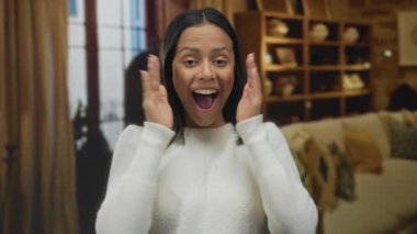 Elleri havada, beyaz süveter giymiş, sıcak bir oturma odasında sevinç ve şaşkınlık içinde gülümseyen bir kadın..