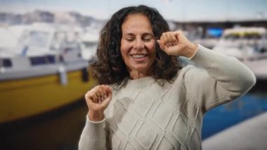 Tekneli bir limanın yanında neşeli, kıvırcık saçlı bir kadın, açık deniz manzarasında rahat bir kazak giyiyor, canlı ve kaygısız bir anı yakalıyor..