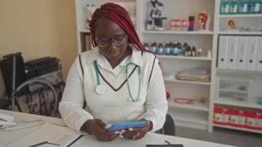 Klinikte steteskopla çalışan bir kadın sağlık ekipmanları ve tıp kitaplarıyla çevrili bir klinikteki cihazdan okuma yapıyor.