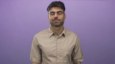 Gözlüklü ve bej gömlekli İspanyol genç adam mor stüdyoda göğsünü yumrukluyor, samimiyet..