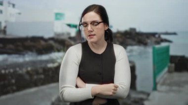 Okyanus zemininde kayalık kıyıda duran kadın, gündüz kumsal ortamında gözlük takarak sakin bir güven yayıyor, sakin deniz manzarasına tezat oluşturuyor..