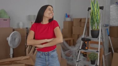 Yeni bir evde neşe içinde dans eden bir kadın. Parlak oturma odasında kutular ve merdivenlerle çevrili. Yeni bir başlangıç için heyecan ve mutluluğu simgeliyor..